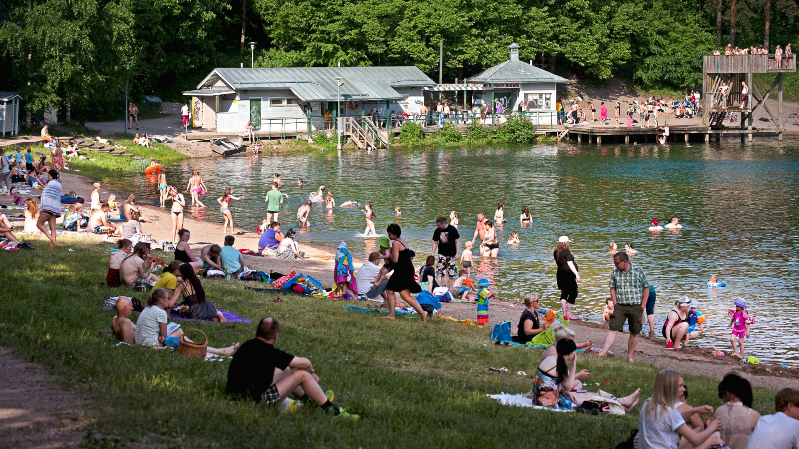 Ihmisiä viettämässä kesäpäivää Mytäjäisten rannalla. Taustalla näkyy pukuhuone, laituri ja hyppytori.