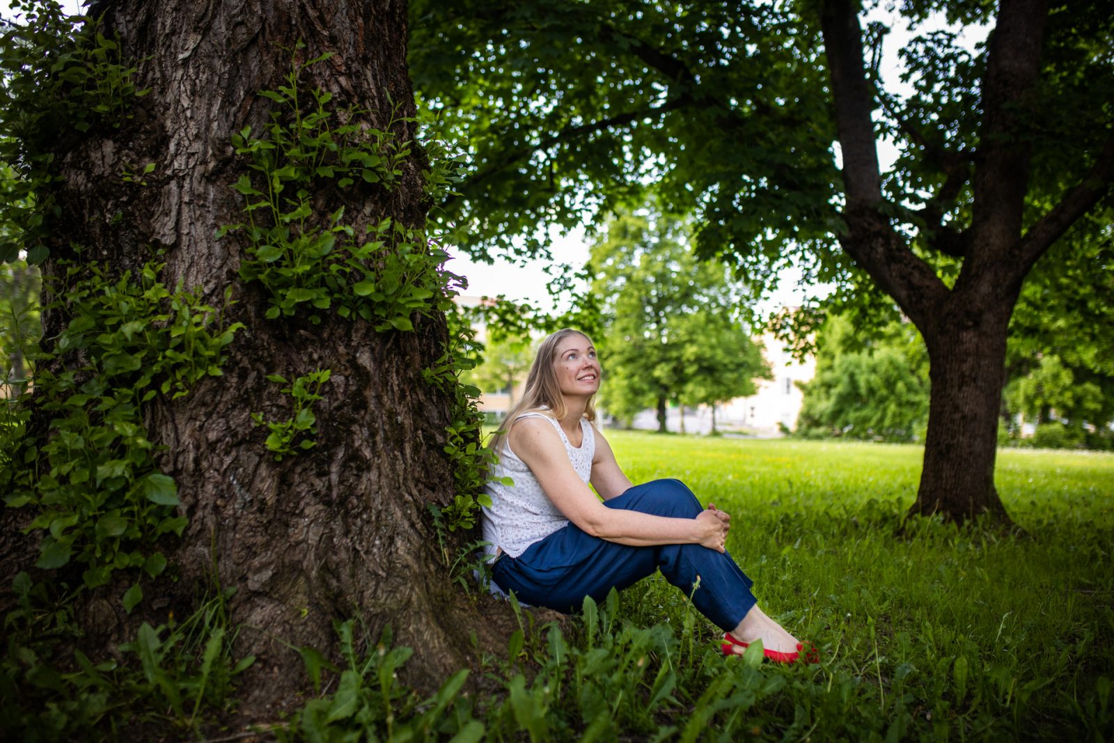 Henkilö istuu puun juurella nurmikolla.