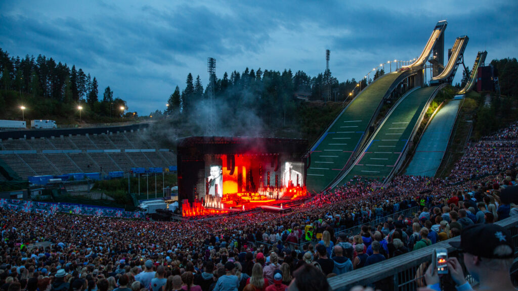 Yleisömassa ison konserttilavan edessä. Taustalla kolme mäkihyppymäkeä.