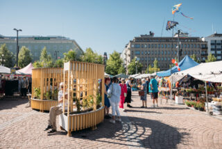 Lahden kauppatorilla on kesäisenä päivänä ihmisiä. Edustalla on puinen penkki, jossa on erilaisia istutuksia