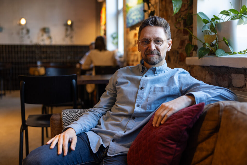 Mies istuu rennosti sohvalla käsi selkänojalla ja katsoo kameraan.