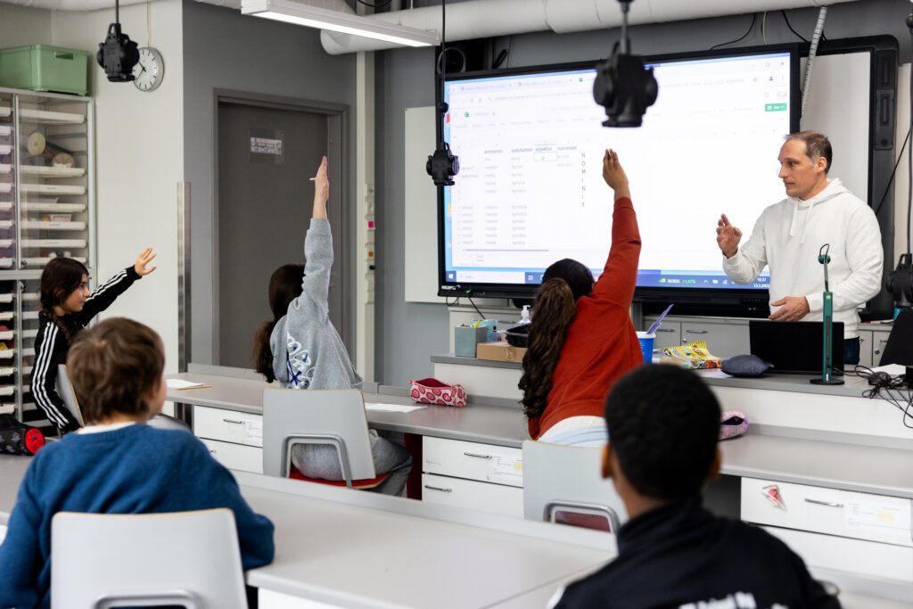 Esa Liukkonen seisoo luokan edessä. Luokassa on viisi oppilasta, joista kolme viittaa. Kuva on otettu oppilaisen selkien takaa, opettajan kasvot näkyvät.