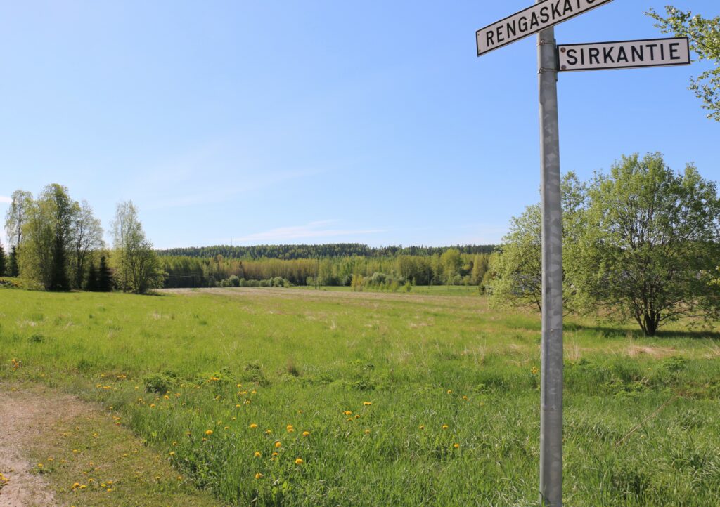 Kuvassa peltoa ja kaukana takana siintää metsänreuna. Sininen taivas, vihreä pelto. Etualalla tienviitta Rengaskadun ja Sirkantien risteyksessä.