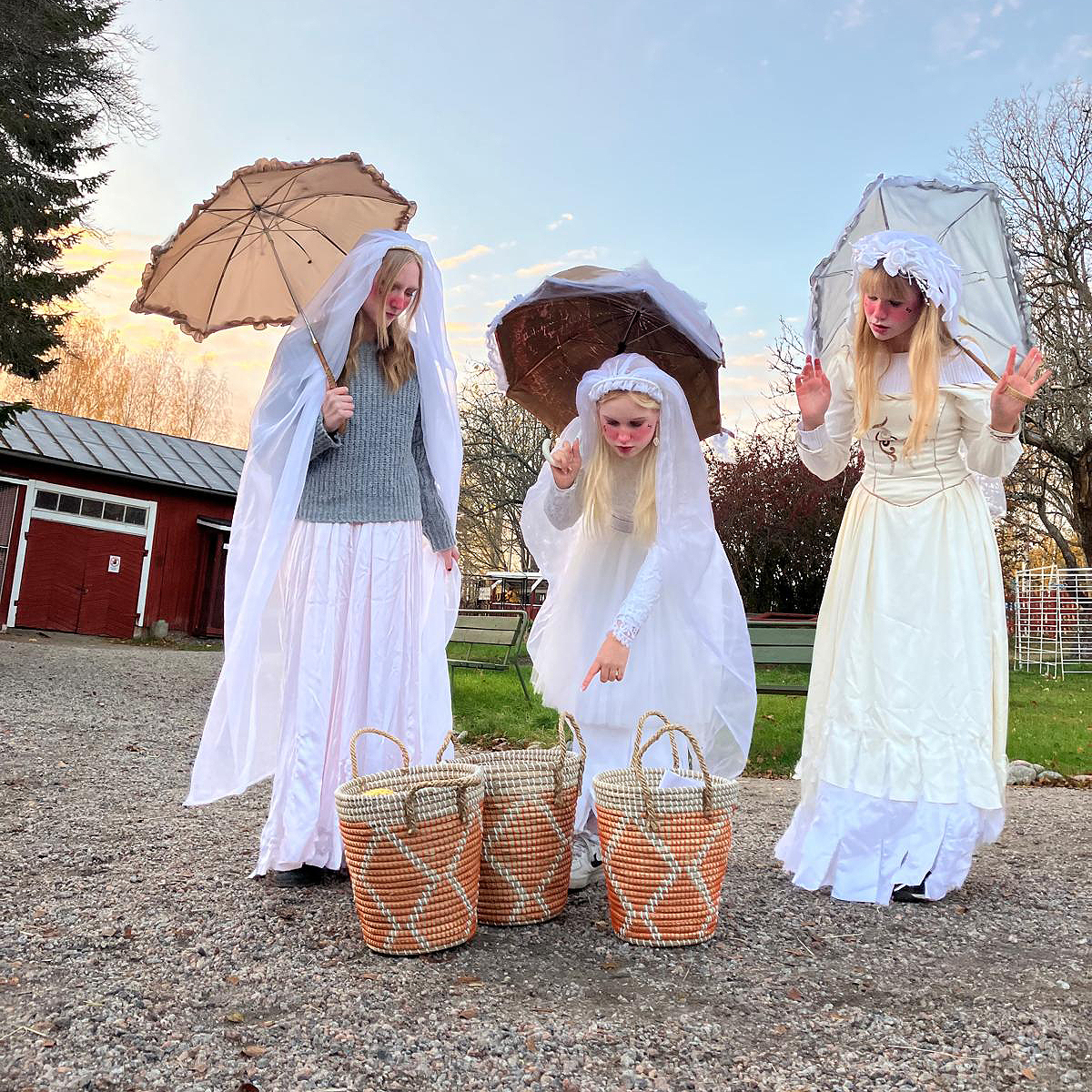 Kolme valkoiseen keijumaiseen asuun pukeutunutta nuorta naista ihmettelee maatilan pihapiirissä maassa olevia koreja. Koreja on kolme.