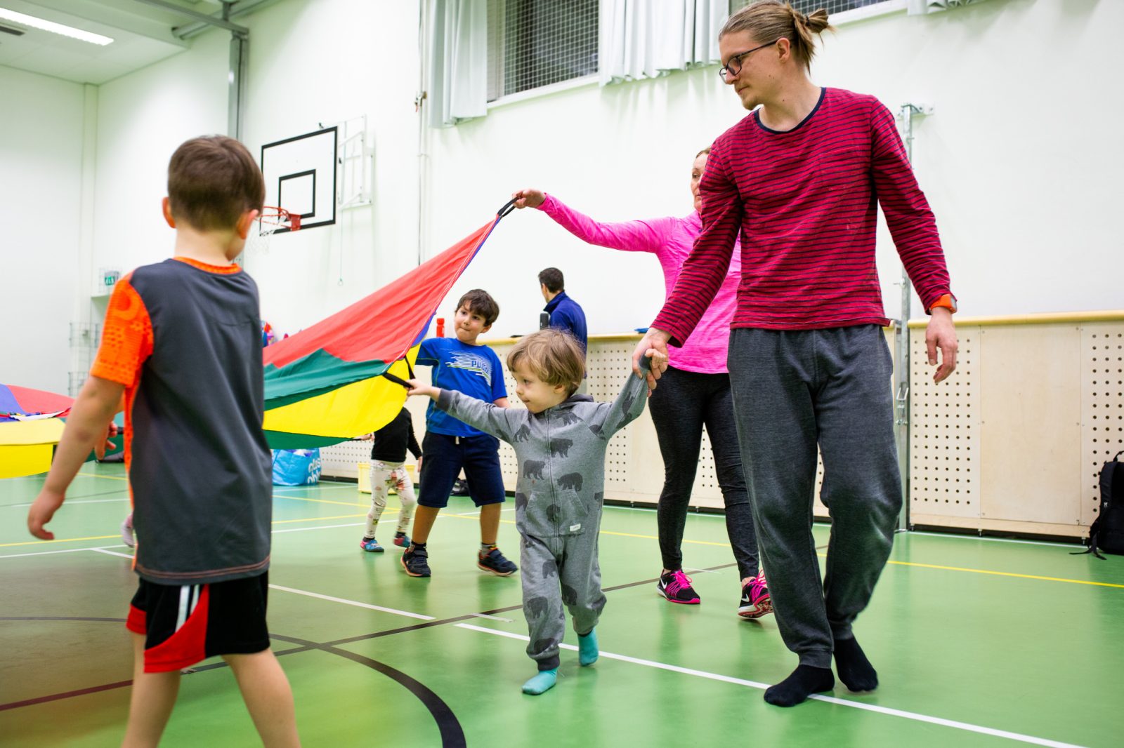 Lapsia ja vanhempia liikkumassa leikkivarjon kanssa liikuntasalissa.