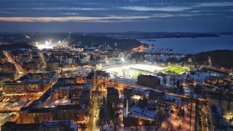 Iltahämäräsää otettu ilmakuva Lahdesta Vesijärven ja Salpausselän suuntaan. Kisapuisto ja urheilukeskus loistavat valoisina kohtina.