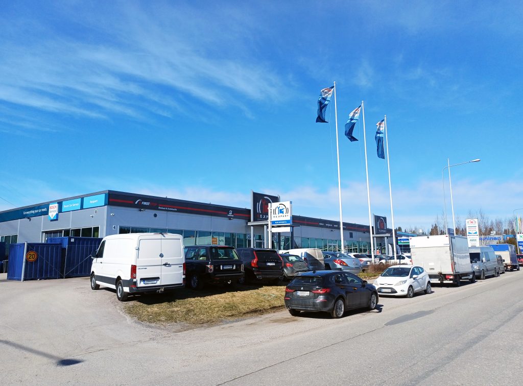 Kadulta otettu kuva autoliikkeestä. Paljon autoja pihassa ja kadunvarressa. Sininen taivas.