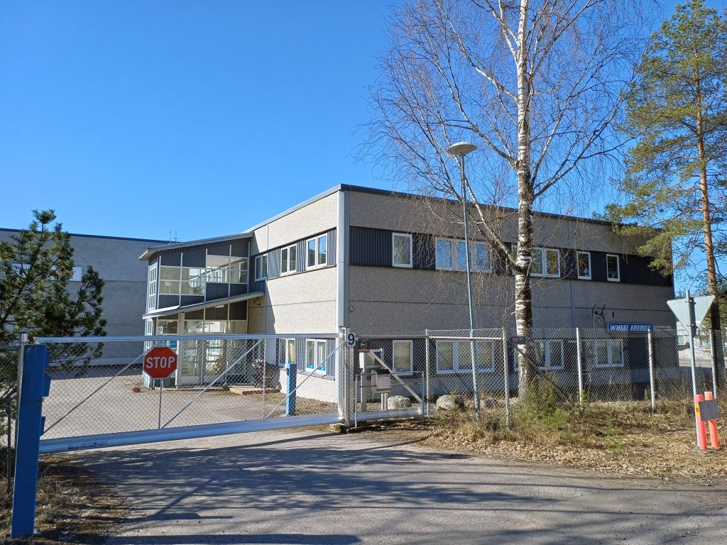 Kuvassa verkkoaidan takan a oleva kaksikerroksinen teollisuuskuunteistö ja puomi kiinni oleva ajoportti.