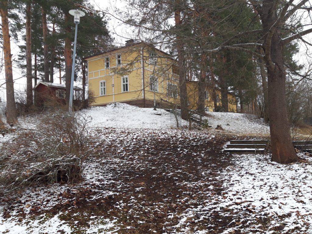 Kuvassa keltainen vanha puinen kaksikerroksinen asuinrakennus. Puiden ympäröimänä pienellä kumpareella.