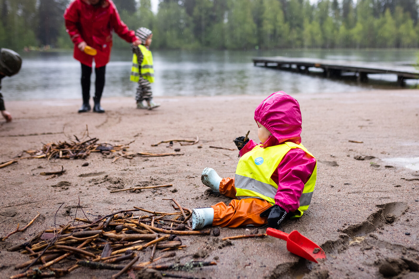 Pieni lapsi istuu lammen rannalla hiekalla ja tutkii lyhyttä puutikkua. Tausta aikuisia.