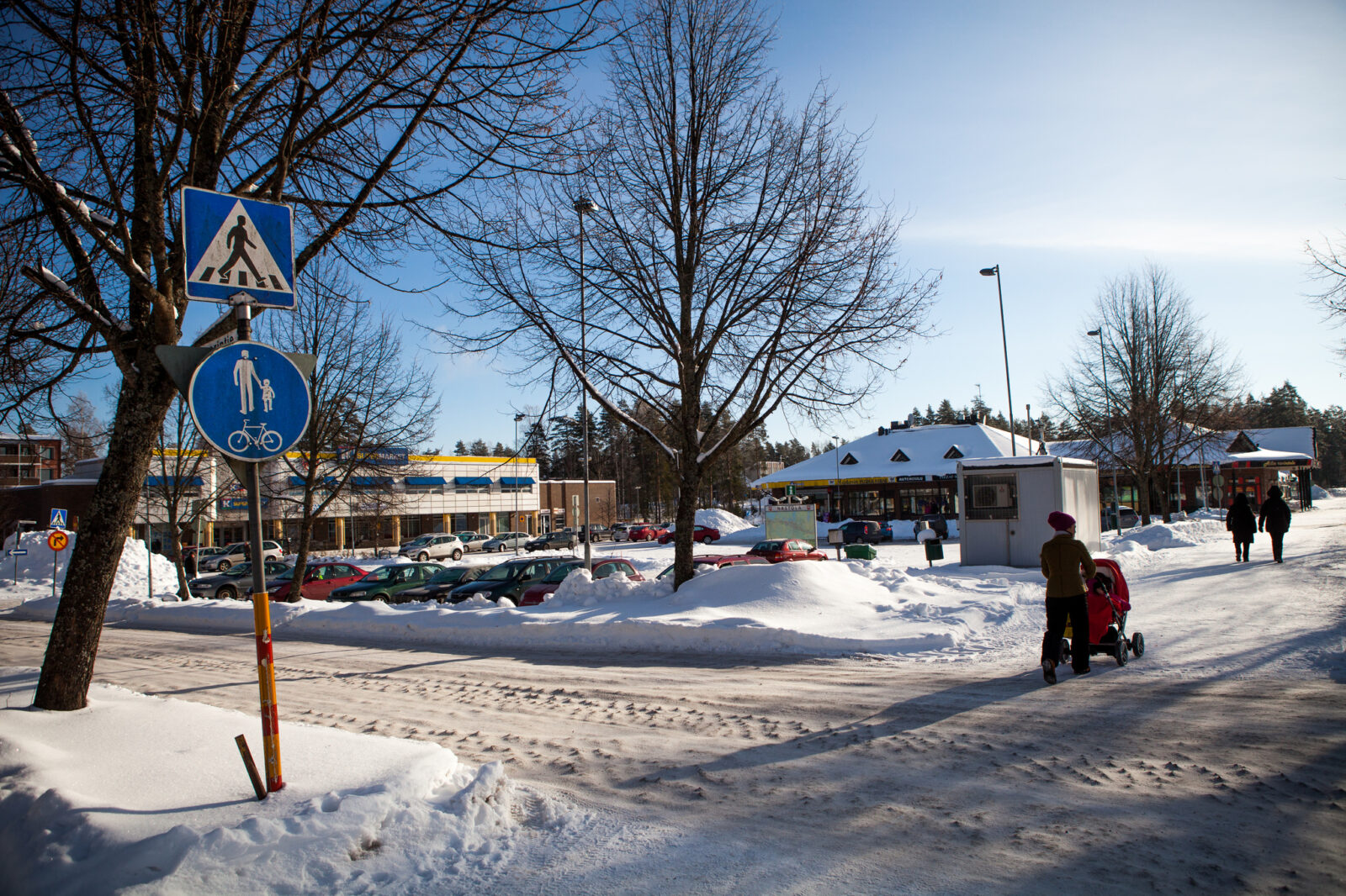 Nainen työntää lastenvaunuja talvisessa maisemassa. Taustalla näkyy Rakokiven ostoskeskus.