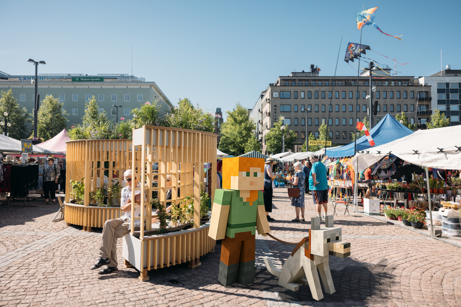 Kuvassa on Lahden kauppatori vilkkaana kesäisenä päivänä. Torilla on puinen penkki istutuksineen ja taustalla on torikojuja ja ihmisiä viettämässä aikaa Lahden kauppatorilla. Kuvassa on myös Minecraft-pelin hahmo, joka on tupsahtanut pelimaalmasta Lahden torille yhdessä koiransa kanssa.
