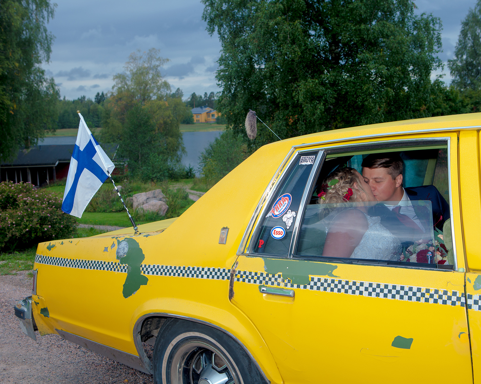Sepposen valokuva pariskunnasta suutelemassa kirkkaankeltaisessa autossa, jonka perässä liehuu Suomen lippu.