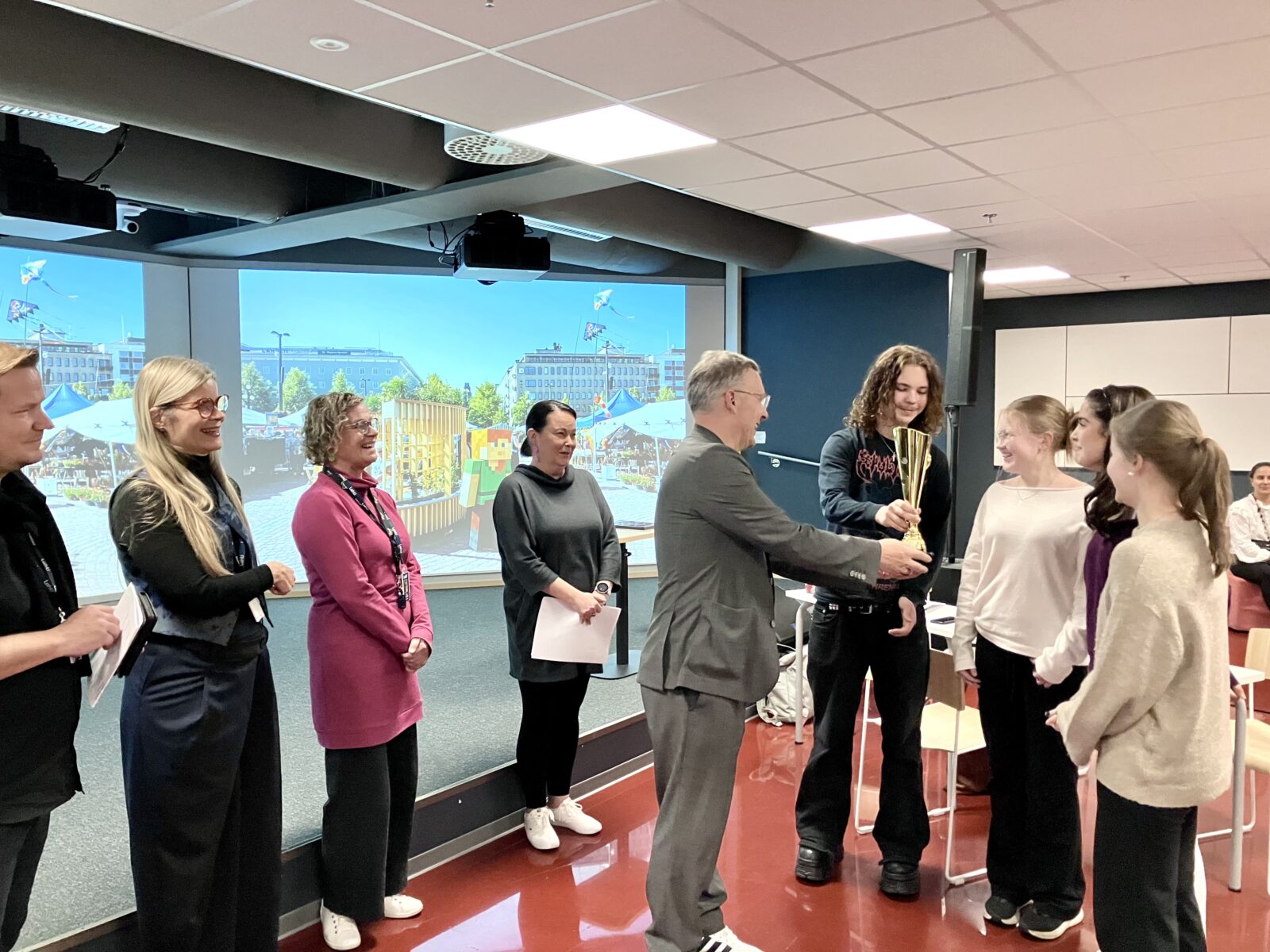 Lahden kaupunginjohtaja ojentaa Minethonin voittajajoukkueelle Ahtialan Ahkerimmille pokaalin parhaasta suunnitelmasta Lahden tulevaisuuden kauppatoriksi. Raati on mukana onnittelemassa.