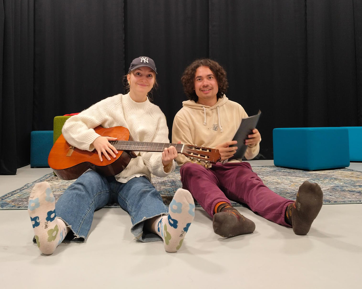 Nuori nainen ja nuori mies istuvat lattialla. Naisella on kitara ja miehellä vihkonen. Taustalla näkyy musta esirippu ja sinisiä raheja.
